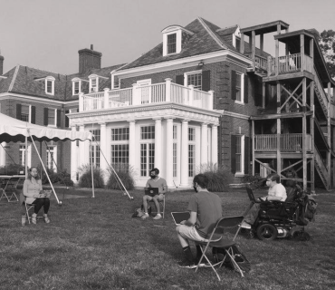 Students sit outside on a lawn outside of a large house. They are on folding chairs set 6 feet apart from each other and on their laptops.